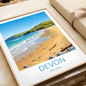 Puede incluir: Una impresión enmarcada que presenta una escena de playa en Devon, Inglaterra. La obra de arte representa una playa de arena con agua turquesa, colinas verdes y un cielo azul claro. El texto "DEVON ENGLAND" está impreso en azul en la parte inferior.