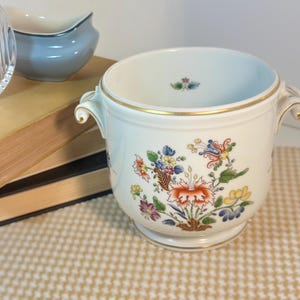 May include: A white porcelain sugar bowl featuring a colourful floral pattern with orange, blue, yellow, and green petals. The bowl has two curved handles, a gold-trimmed rim, and a small floral motif painted on the interior base.