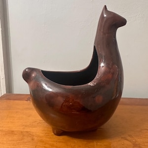 May include: A brown ceramic llama-shaped planter with a glossy finish. The planter has a rounded body, a long neck, and a small opening for plants. It is sitting on a wooden surface.