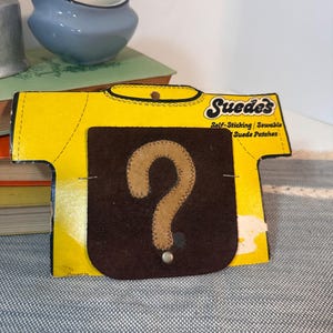 May include: A dark brown square suede patch featuring a tan question mark appliqué, mounted on a yellow t-shirt shaped cardboard backing labeled Suede's Self-Sticking Sewable Suede Patches with a small metal stud at the bottom of the patch.