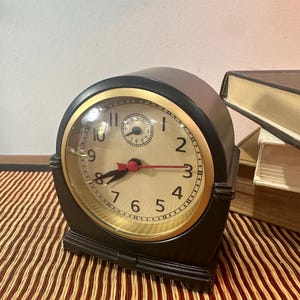 May include: A black mantel clock with a cream-colored face, gold-toned bezel, and black Arabic numerals. The clock features black hour and minute hands, a red second hand, and a small circular sub-dial at the top center of the clock face.