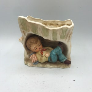 May include: A ceramic figurine of a boy sleeping in a tree trunk with a red and white accordion. The figurine is painted in shades of brown, green, blue, and white.