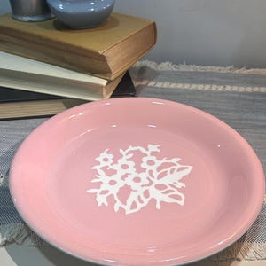Pode incluir: Um prato de cerâmica rosa com um desenho floral branco no centro. O prato é redondo com uma borda ligeiramente elevada. Outros itens no fundo incluem um jarro de metal, uma pequena tigela azul e uma pilha de livros.
