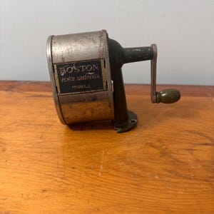 May include: A vintage Boston pencil sharpener, Model J, with a silver metal body and a dark green handle. The text "BOSTON PENCIL SHARPENER" is visible on the front. The sharpener is set on a wooden surface.