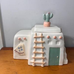 Pode incluir: Um pote de biscoitos de cerâmica em forma de edifício adobe do sudoeste. O pote é branco sujo com uma porta verde, uma escada castanha e um cacto no topo. Elementos decorativos: pequenos círculos cor-de-rosa e um padrão.