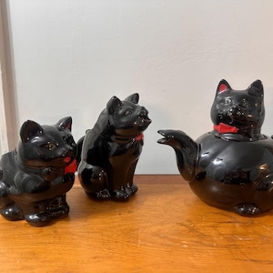 May include: A black ceramic cat-themed tea set, comprising a teapot and two matching figurines. The teapot and figurines have red bow ties. The set is displayed on a wooden surface.