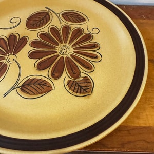 以下が含まれることがあります： 茶色の縁取りのある、大きく丸いベージュのセラミック製の皿。皿には、茶色の花と葉のスタイリッシュな花柄が描かれている。