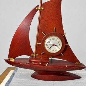 May include: A sailboat-shaped table clock crafted from dark reddish-brown wood. The clock face is circular with a gold-toned rim, black numerals, and a ship's wheel design. It features black hour and minute hands with a red second hand.