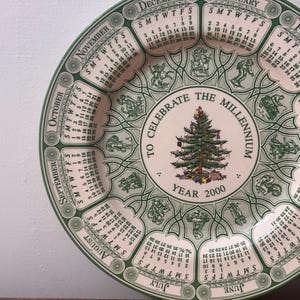 May include: A decorative plate with a calendar design for the year 2000. The plate features a green and white color scheme, with each month's calendar displayed around the edge. A Christmas tree is in the center, with the text "TO CELEBRATE THE MILLENNIUM".