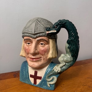 May include: A ceramic mug shaped like a medieval knight's head, featuring a helmet and a dragon-shaped handle. The knight has a stern expression, with a red cross on his blue tunic. The dragon handle is dark green.