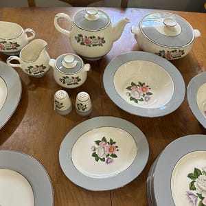 May include: A full vintage porcelain dinner service with a floral pattern. The set includes a teapot, serving bowl, plates, cups, a creamer, sugar bowl, and salt and pepper shakers. The pieces have a light blue rim and a floral pattern with pink and purple flowers.