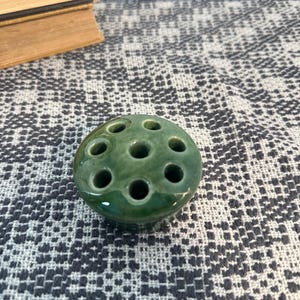 May include: A round, dark green ceramic flower frog featuring a glossy finish and seven circular holes arranged in a pattern on its top surface, designed to hold individual flower stems upright in a floral arrangement.