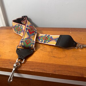May include: A patterned camera strap with a yellow background and geometric designs in red, green, and blue. The strap has black leather ends and metal clips for attachment. The strap is lying on a wooden surface.
