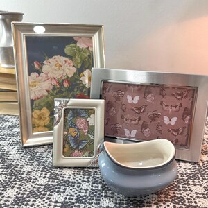 May include: A collection of decorative items, including three silver-framed pictures with floral and butterfly prints, and a small, light blue ceramic bowl. The frames vary in size and shape, creating a layered display.