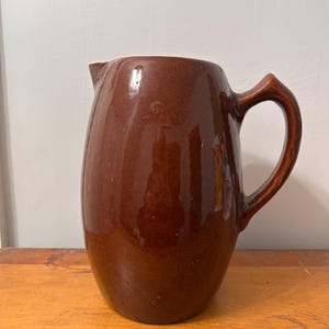 May include: A brown ceramic pitcher with a glossy finish. The pitcher has a rounded body, a curved handle, and a spout for pouring. The pitcher is likely used for serving beverages.