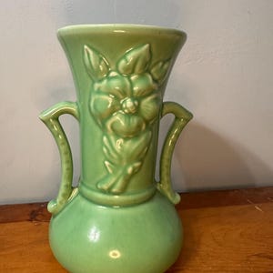 Könnte beinhalten: Eine grüne Keramikvase mit bauchigem Boden und ausgestelltem Oberteil. Die Vase hat zwei gebogene Griffe und ein erhabenes Blumendesign auf der Vorderseite. Die Vase ist etwa 20 cm hoch.