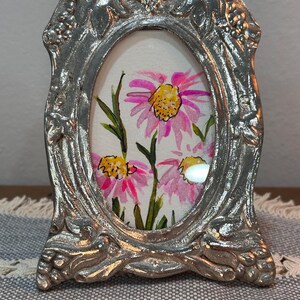 May include: Silver-colored, ornate oval picture frame with a watercolor painting of pink and yellow flowers. The frame has a detailed, textured design. The artwork features pink petals and yellow centers, with green stems and leaves.