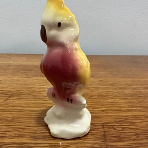 May include: A ceramic figurine of a yellow and pink cockatoo perched on a branch. The bird has a white head and a black eye.