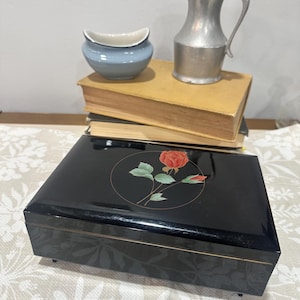 May include: A black rectangular jewelry box with a rose design, a small blue ceramic dish, and a silver metal pitcher are displayed on a stack of books. The jewelry box has a glossy finish and gold trim.