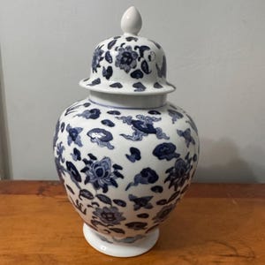 May include: A white and blue ceramic jar with a lid. The jar features a floral and cloud pattern in blue against a white background. The jar is on a wooden surface.