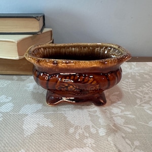 May include: A small, oval-shaped ceramic planter with a glossy, dark brown glaze. The vessel features a raised, textured relief pattern along the sides and rests on four short, sturdy feet. The rim has a lighter, dripped brown glaze effect.