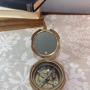 May include: A circular brass pocket compass with a hinged lid featuring an interior mirror. The compass dial displays cardinal directions and numerical degree markings, with a needle pointing toward the north and south indicators.