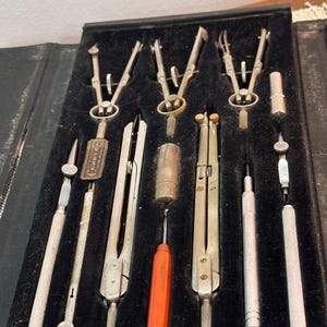 May include: A set of vintage drafting tools, including compasses, dividers, and a screwdriver, arranged in a black case with a velvet lining. The tools are silver-toned with some having a brass or orange handle.