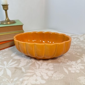 May include: A glossy, pumpkin-orange ceramic bowl with a scalloped, fluted edge and a rounded, shallow basin. The bowl features a smooth, reflective finish and a distinct petal-like design around its exterior circumference.