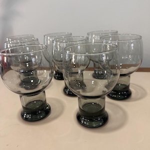 May include: A collection of eight clear, stemless glasses. Each glass has a rounded bowl and a dark grey base. The glasses are grouped together, highlighting their distinctive shape. The glassware is suitable for serving drinks.