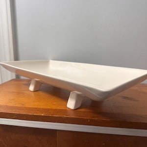 Puede incluir: Una fuente rectangular de cerámica blanquecina con base elevada. La fuente tiene un diseño elegante y moderno y se eleva mediante cuatro pequeños pies cuadrados. La fuente está sobre una superficie de madera.