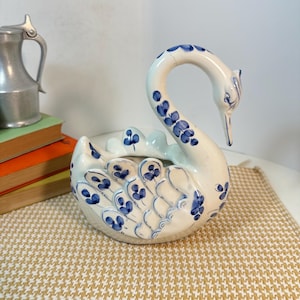 May include: A white ceramic swan-shaped planter or decorative bowl featuring hand-painted blue floral patterns on its wings, neck, and head. The swan has a curved neck and detailed feather motifs, with a hollow center for holding small items.