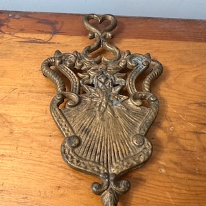 May include: Ornate brass trivet with a heart-shaped handle and intricate floral and scrollwork design. The trivet has a shield-like shape with a sunburst pattern in the center. The brass has a vintage patina.