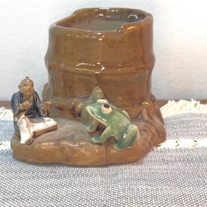 May include: A ceramic decorative vase with a brown, textured exterior. A small figurine of a seated man and a green frog are on the base. The vase has a cylindrical shape with a smooth interior.