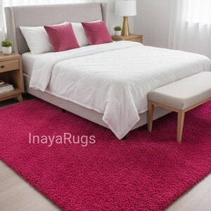 May include: A bedroom scene featuring a plush, magenta rug. The rug is placed in front of a bed with white bedding and magenta pillows. A bench sits at the foot of the bed. The text "InayaRugs" is visible on the rug.