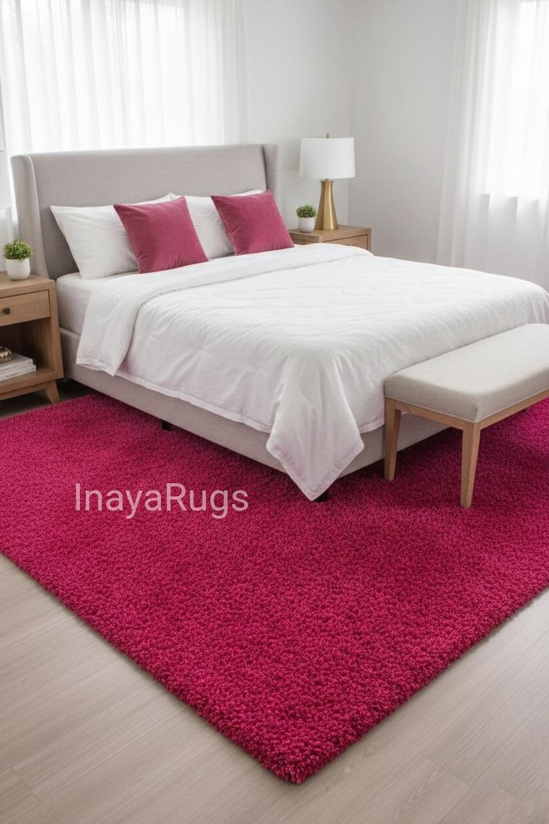 May include: A bedroom scene featuring a plush, magenta rug. The rug is placed in front of a bed with white bedding and magenta pillows. A bench sits at the foot of the bed. The text "InayaRugs" is visible on the rug.