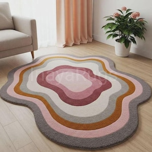 Könnte beinhalten: Ein einzigartiger, abstrakt geformter Teppich mit einem geschichteten Design. Der Teppich hat einen wellenförmigen Umriss und eine Farbpalette aus Grau, Rosa, Weiß und Gold. Der Teppich liegt auf einem Holzboden, mit einem neutralen Sessel und einer Topfpflanze im Hintergrund.