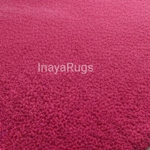 May include: A close-up of a textured, raspberry-colored rug. The rug's surface is covered in small, tightly packed loops, creating a plush appearance. The text "InayaRugs" is printed in white across the rug's surface.