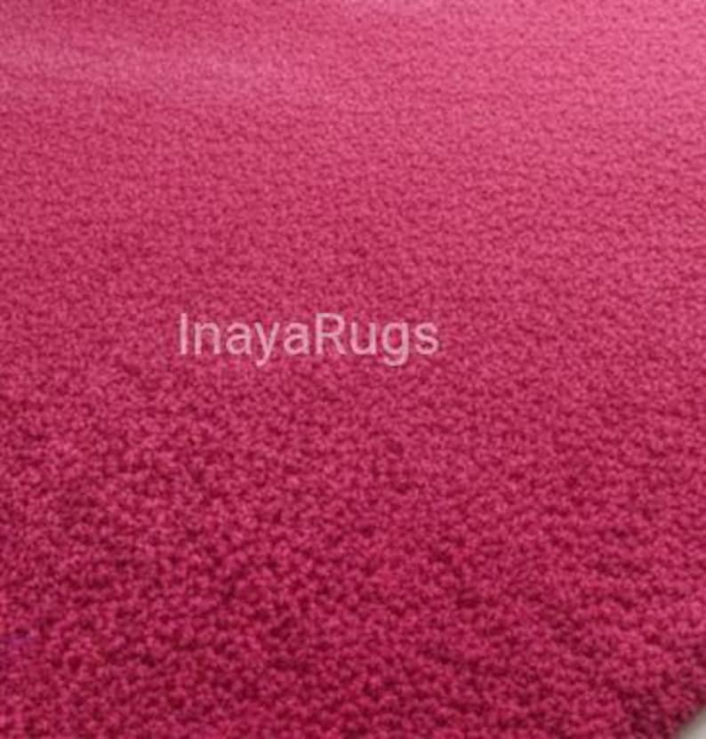 May include: A close-up of a textured, raspberry-colored rug. The rug's surface is covered in small, tightly packed loops, creating a plush appearance. The text "InayaRugs" is printed in white across the rug's surface.
