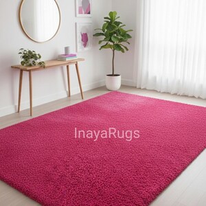 May include: A large, rectangular, magenta-colored shag rug. The rug is the focal point of a room with light-colored wood flooring, white walls, and a sheer white curtain. The text "InayaRugs" is visible on the rug.