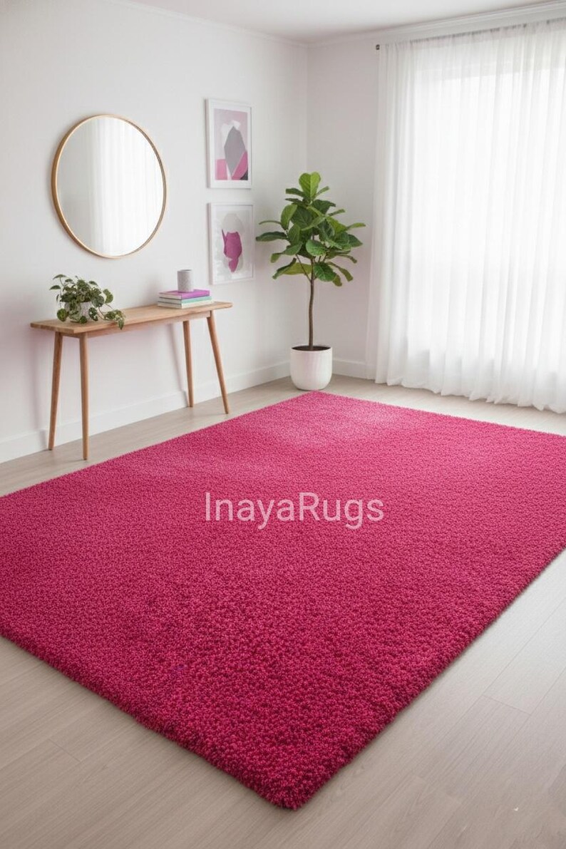 May include: A large, rectangular, magenta-colored shag rug. The rug is the focal point of a room with light-colored wood flooring, white walls, and a sheer white curtain. The text "InayaRugs" is visible on the rug.