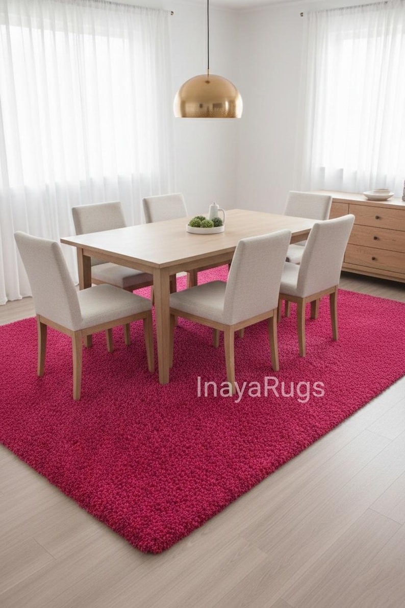 May include: A dining room set with a light wood table and six beige upholstered chairs. The room features a vibrant fuchsia rug, a gold pendant light, and sheer white curtains. A wooden sideboard is visible in the background.