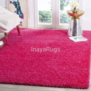 May include: A vibrant pink rectangular shag rug. The rug has a plush, textured surface and covers most of the floor. The text "InayaRugs" is visible on the rug. A white chair and a decorative table are in the background.