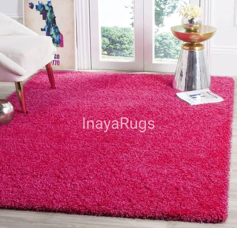 May include: A vibrant pink rectangular shag rug. The rug has a plush, textured surface and covers most of the floor. The text "InayaRugs" is visible on the rug. A white chair and a decorative table are in the background.