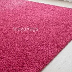 May include: A rectangular, plush, fuchsia-colored rug. The rug has a dense, textured surface and is placed on a light-colored wooden floor. The text "InayaRugs" is visible on the rug.
