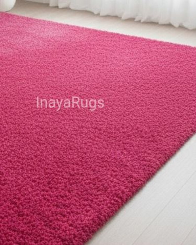 May include: A rectangular, plush, fuchsia-colored rug. The rug has a dense, textured surface and is placed on a light-colored wooden floor. The text "InayaRugs" is visible on the rug.