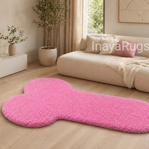 May include: A bright pink, shaggy rug in a unique shape, resembling a phallus. The rug is placed on a light wood floor in a living room setting. The rug's texture appears soft and plush.