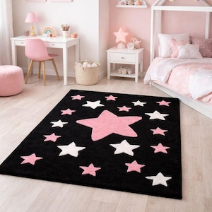 Könnte beinhalten: Schwarzer rechteckiger Teppich mit rosa und weißen Sternen. Der Teppich hat einen großen rosa Stern in der Mitte, umgeben von kleineren rosa und weißen Sternen. Der Teppich befindet sich in einem rosa Schlafzimmer.