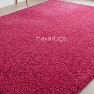 May include: A plush, magenta-colored rug with a textured surface. The rug fills the frame, showcasing its dense, soft pile. The text "InayaRugs" is visible on the rug's surface. The rug is a solid color and appears to be made of a soft material.