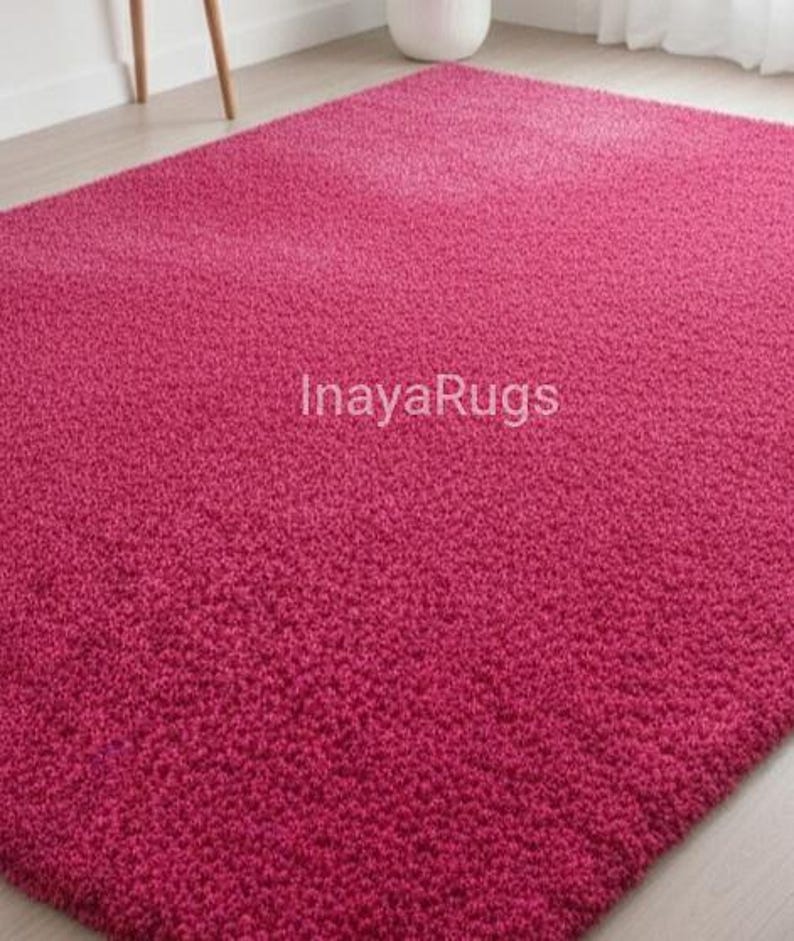May include: A plush, magenta-colored rug with a textured surface. The rug fills the frame, showcasing its dense, soft pile. The text "InayaRugs" is visible on the rug's surface. The rug is a solid color and appears to be made of a soft material.