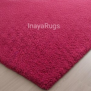 May include: A close-up of a textured, magenta-colored rug. The rug has a dense, plush pile and the text "Inaya Rugs" is visible. The rug's corner is visible, showing its thick construction.